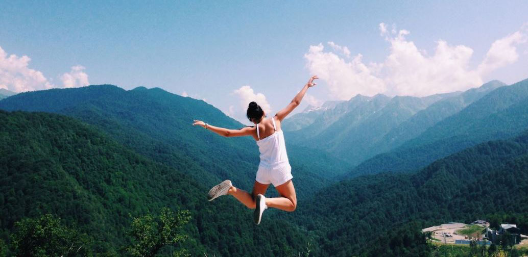 A lady jumping high in the air with her arms out wide above a green mountain valley